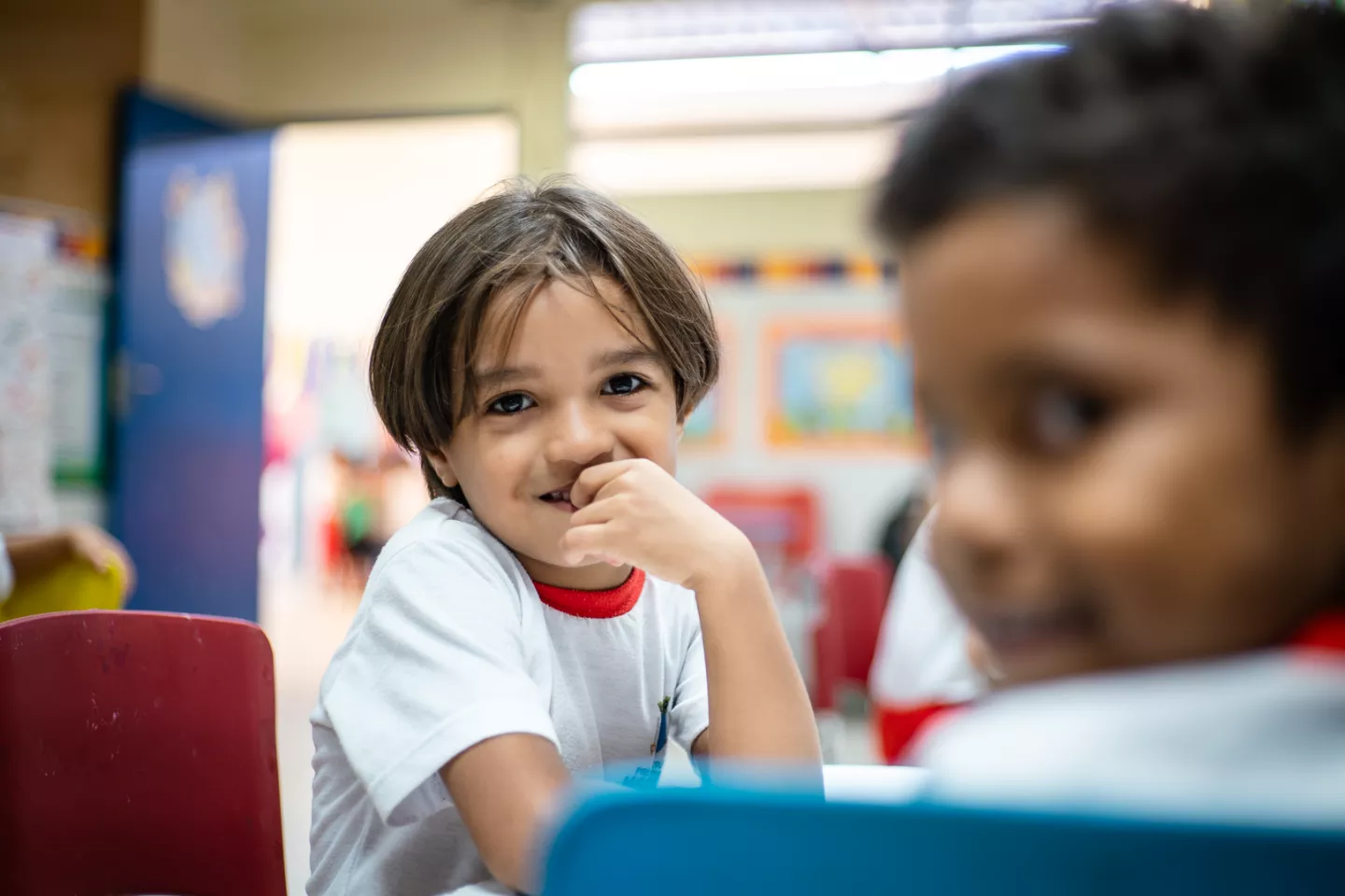 Foto mostra um menino pequeno em uma sala de aula. Ele est&aacute; com a m&atilde;o no rosto, sorrindo. Em primeiro plano na foto, mas desfocado, h&aacute; outro menino sorrindo.
