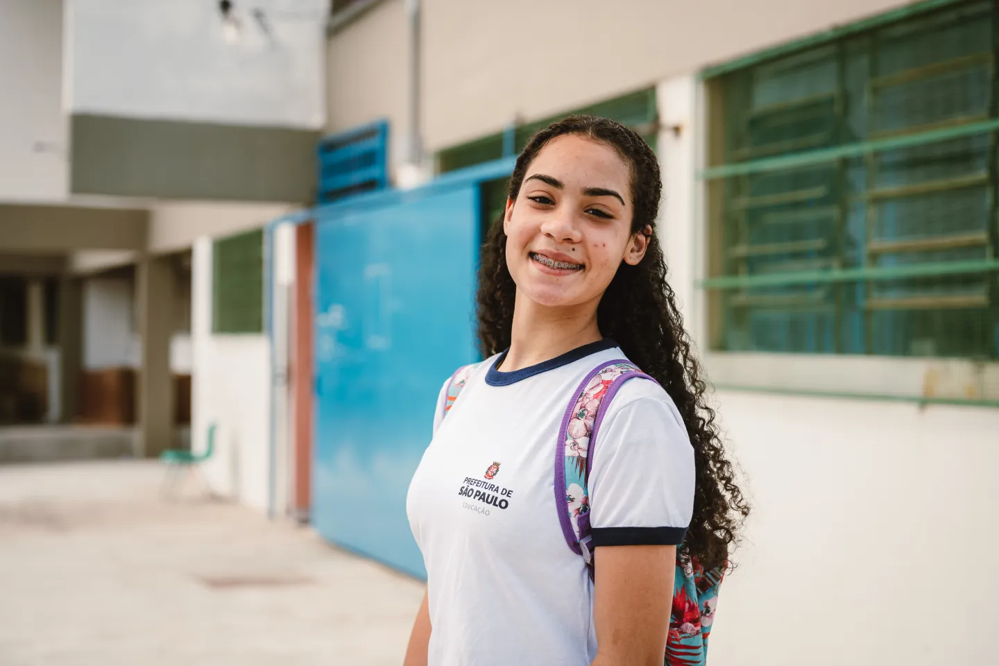 Foto mostra uma menina de uniforme escolar e mochila nas costas, parada o corredor de uma escola. Ela est&aacute; olhando para a c&acirc;mera, sorrindo.