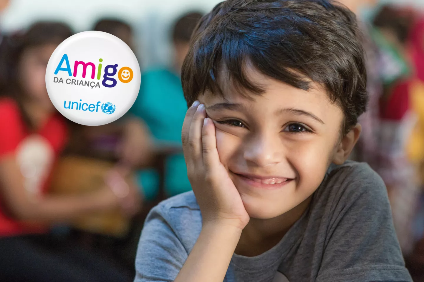 Foto mostra um menino sorridente, apoiando o rosto na m&atilde;o. Em cima da foto, do lado esquerdo, h&aacute; a marca do programa Amigo da Crian&ccedil;a, do UNICEF Brasil