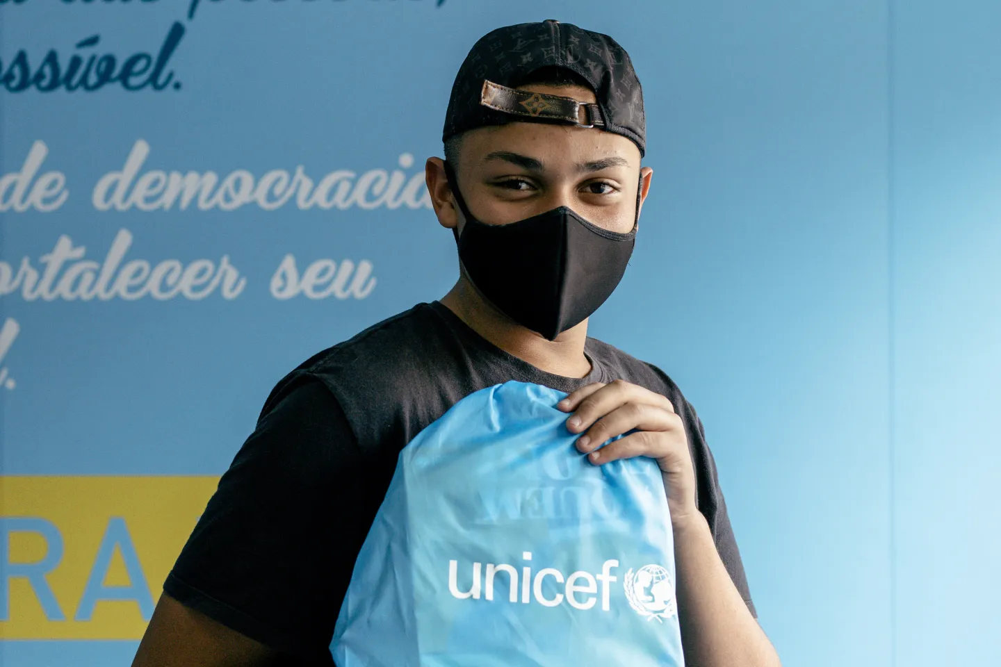 Foto mostra um adolescente em p&eacute; em frente a uma parede. Ele est&aacute; segurando uma mochila azul com o logo do UNICEF. Ele est&aacute; usando um bon&eacute; com a aba virada para tr&aacute;s e m&aacute;scara. Apesar da m&aacute;scara, nota-se que o adolescente est&aacute; sorrindo.