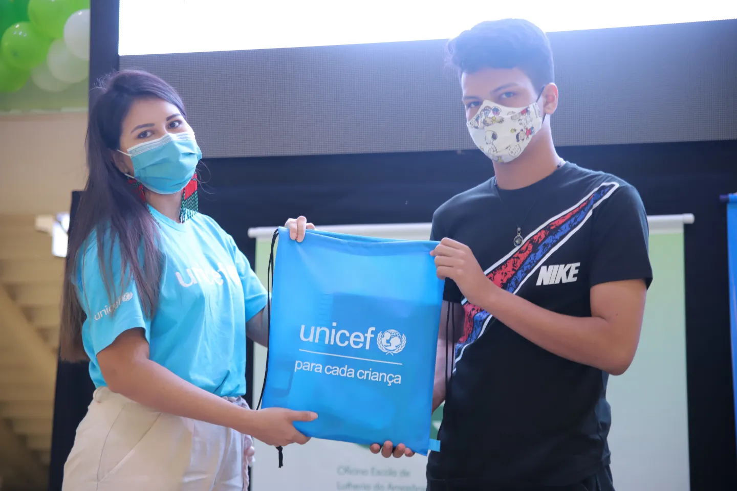 Foto mostra uma profissional com a camiseta do UNICEF entregando uma mochila com o logo do UNICEF para um adolescente. Os dois est&atilde;o de m&aacute;scara e mant&eacute;m dist&acirc;ncia entre eles.