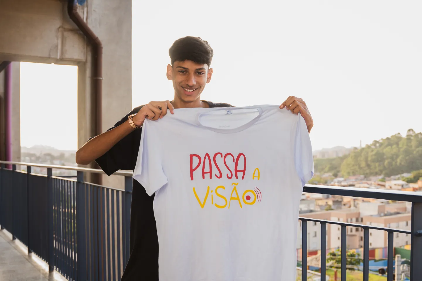 Foto mostra um adolescente segurando uma camiseta com o logo do projeto Passa a Vis&atilde;o. Atr&aacute;s dele h&aacute; constru&ccedil;&otilde;es urbanas.