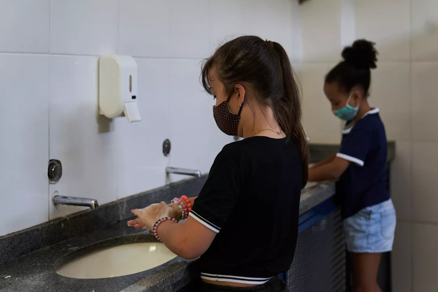 Foto mostra duas meninas lavando as m&atilde;os no banheiro da escola. As duas usam m&aacute;scara e est&atilde;o distantes 1,5 metro uma da outra
