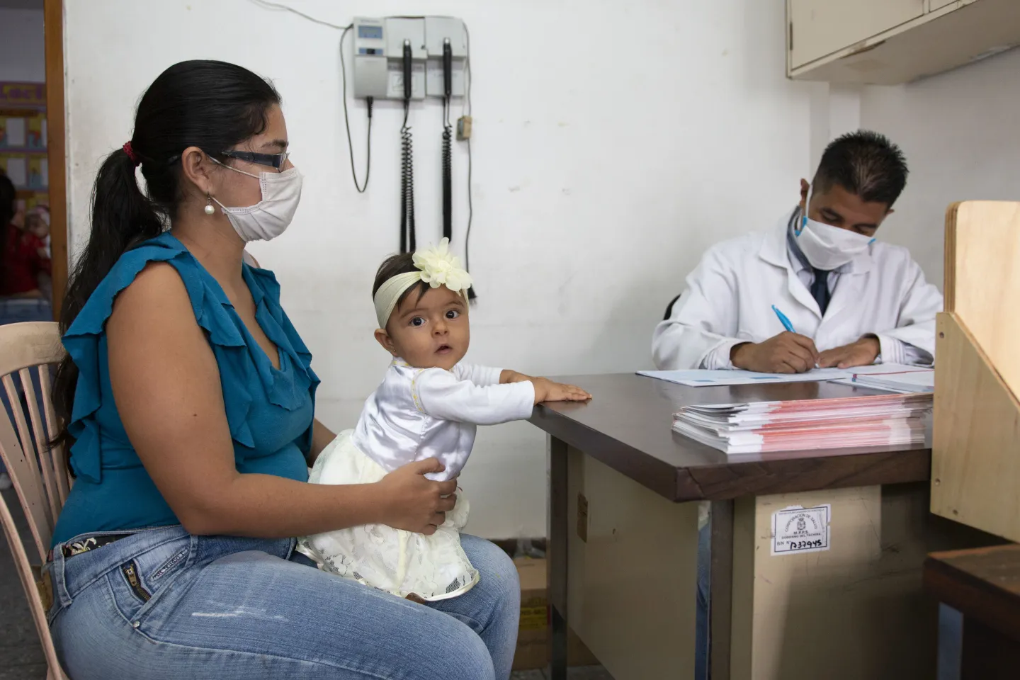 uma m&atilde;e est&aacute; em um consult&oacute;rio m&eacute;dico com sua beb&ecirc;. ela e o m&eacute;dico usam m&aacute;scara. a beb&ecirc; est&aacute; olhando para a c&acirc;mera