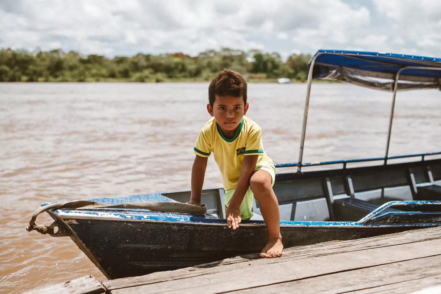 Foto mostra um menino saindo de um barco. Atr&aacute;s dele est&aacute; o Rio Amazonas e ao fundo a floresta.