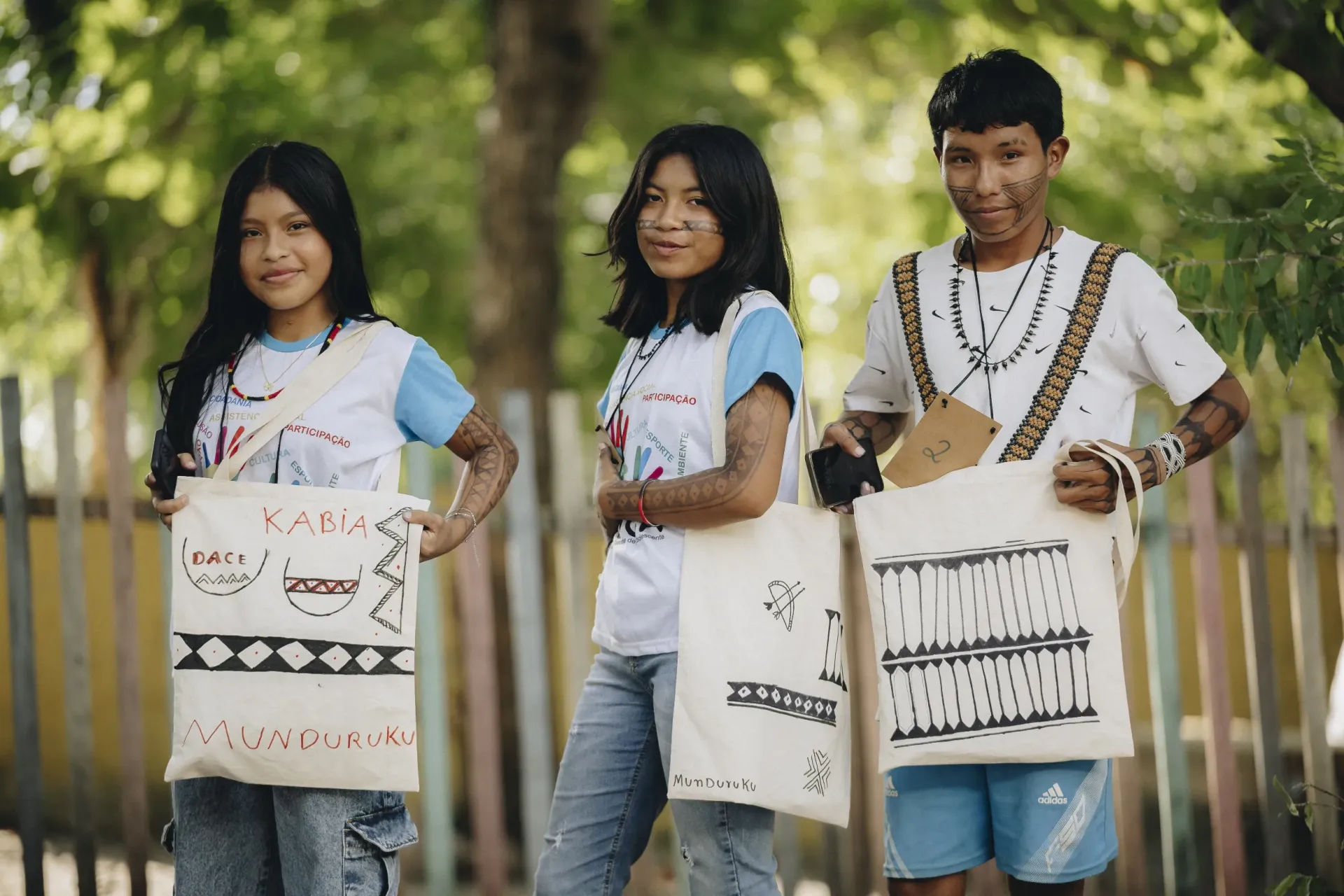 tr&ecirc;s jovens ind&iacute;genas posam para foto segurando bolsas de pano em meio &agrave; floresta