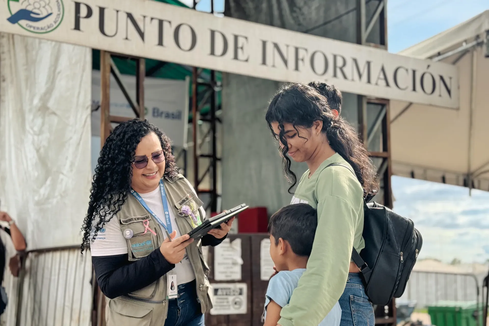 funcion&aacute;ria com colete identificado com UNICEF fala com mulher e crian&ccedil;a em frente a um Punto de Informa&ccedil;&atilde;o