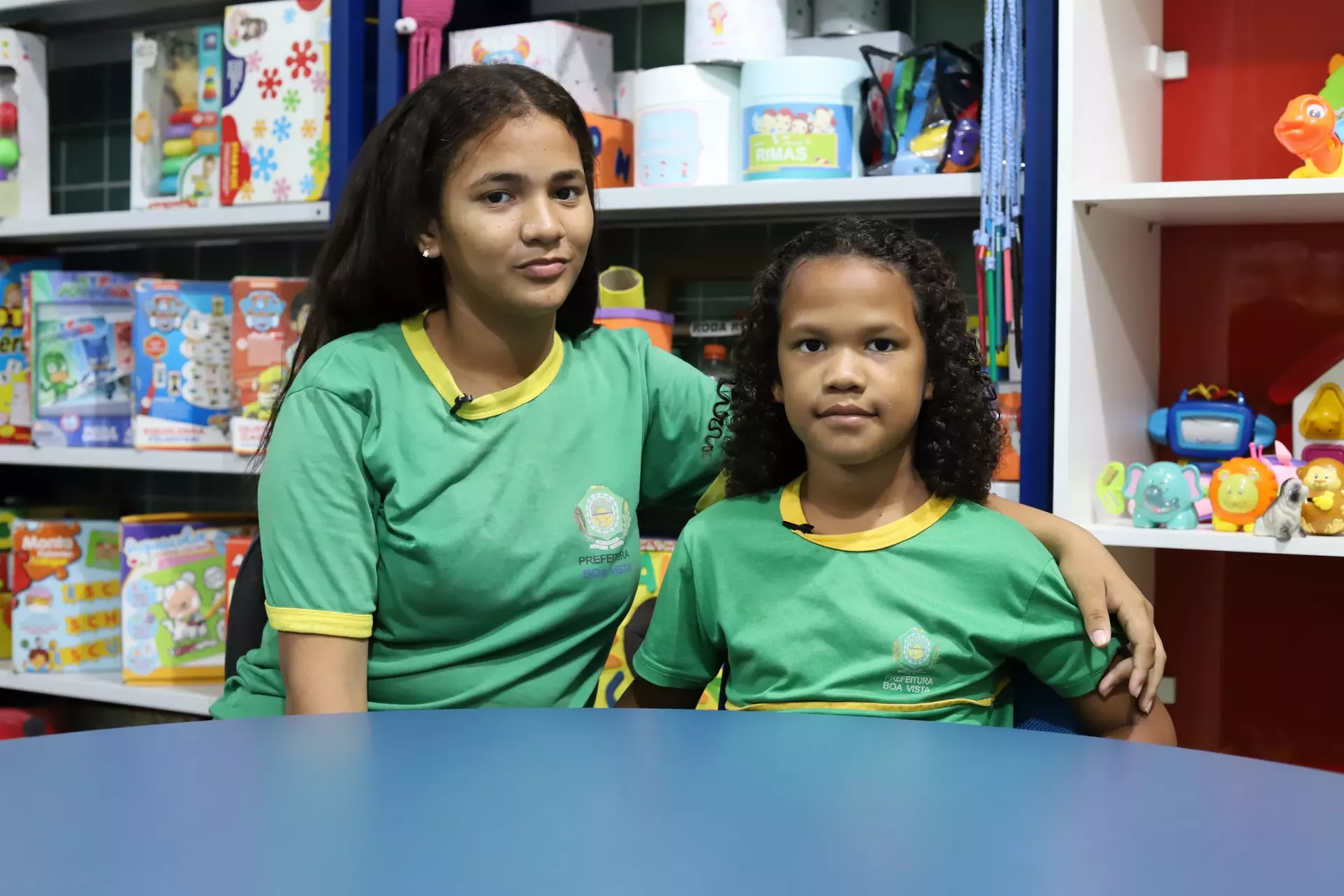 duas estudantes se abra&ccedil;am enquanto sentadas em uma mesa de biblioteca escolar