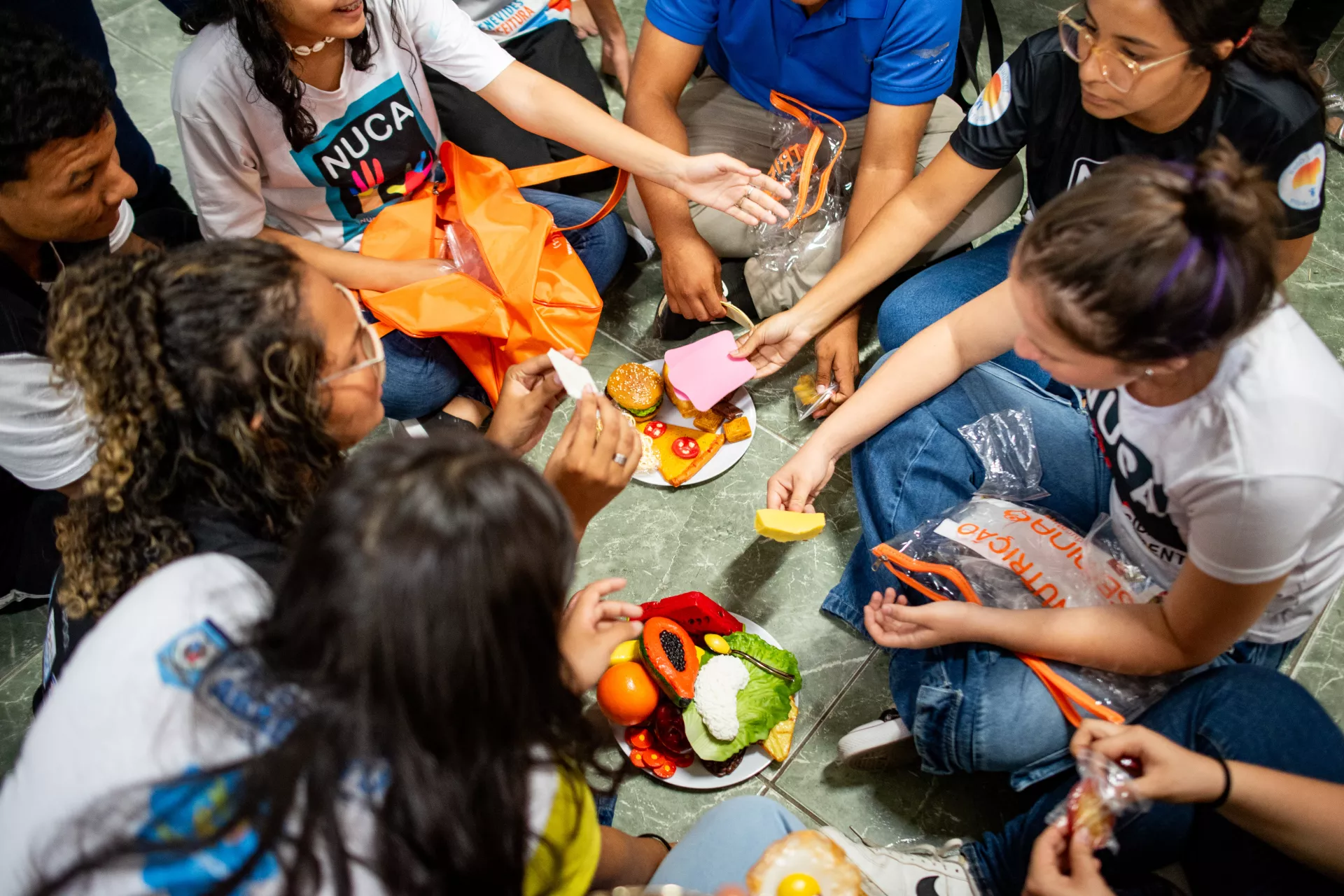 Foto mostra v&aacute;rios adolescentes sentados no ch&atilde;o em volta de pratos de comida