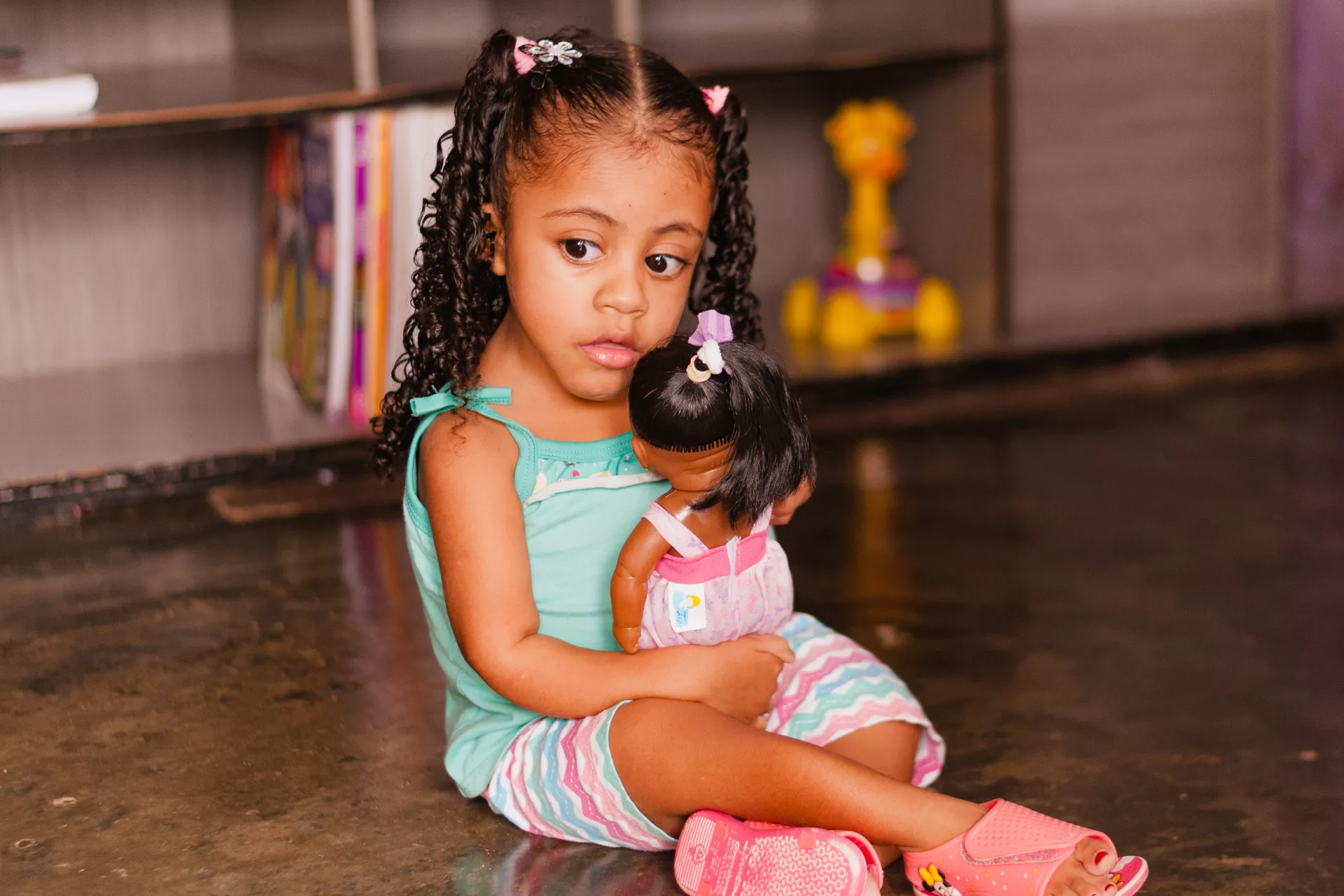 Foto mostra uma menina pequena sentada com uma boneca no colo.