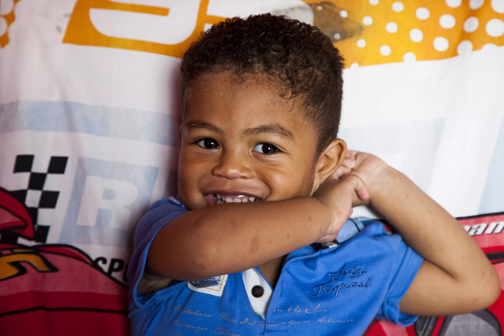 um menino pequeno olha para a c&acirc;mera sorrindo, ele est&aacute; deitado em um travesseiro