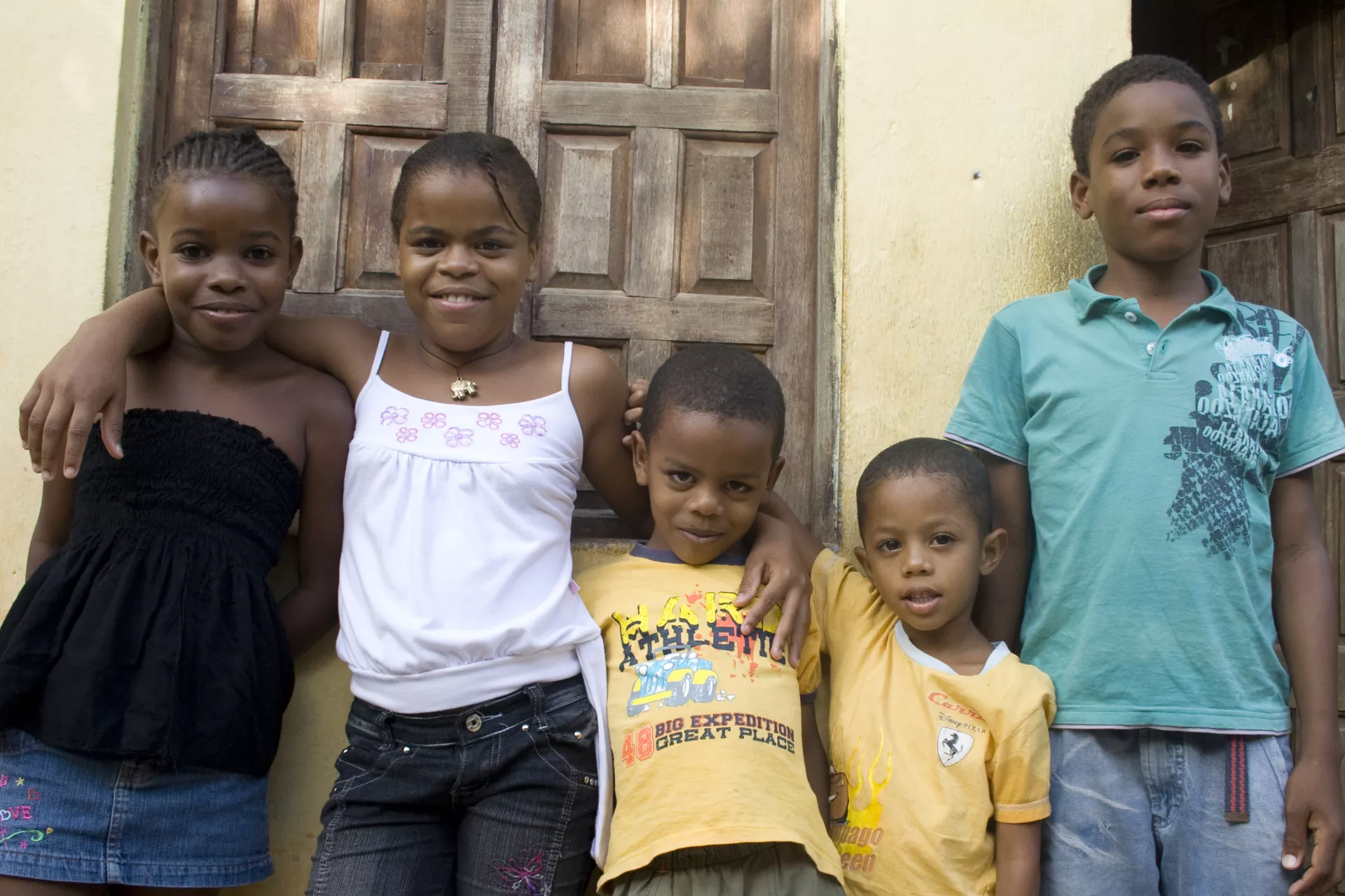 Cinco crian&ccedil;as de diferentes idades est&atilde;o encostadas em uma parede externa. Elas olham para a c&acirc;mera.