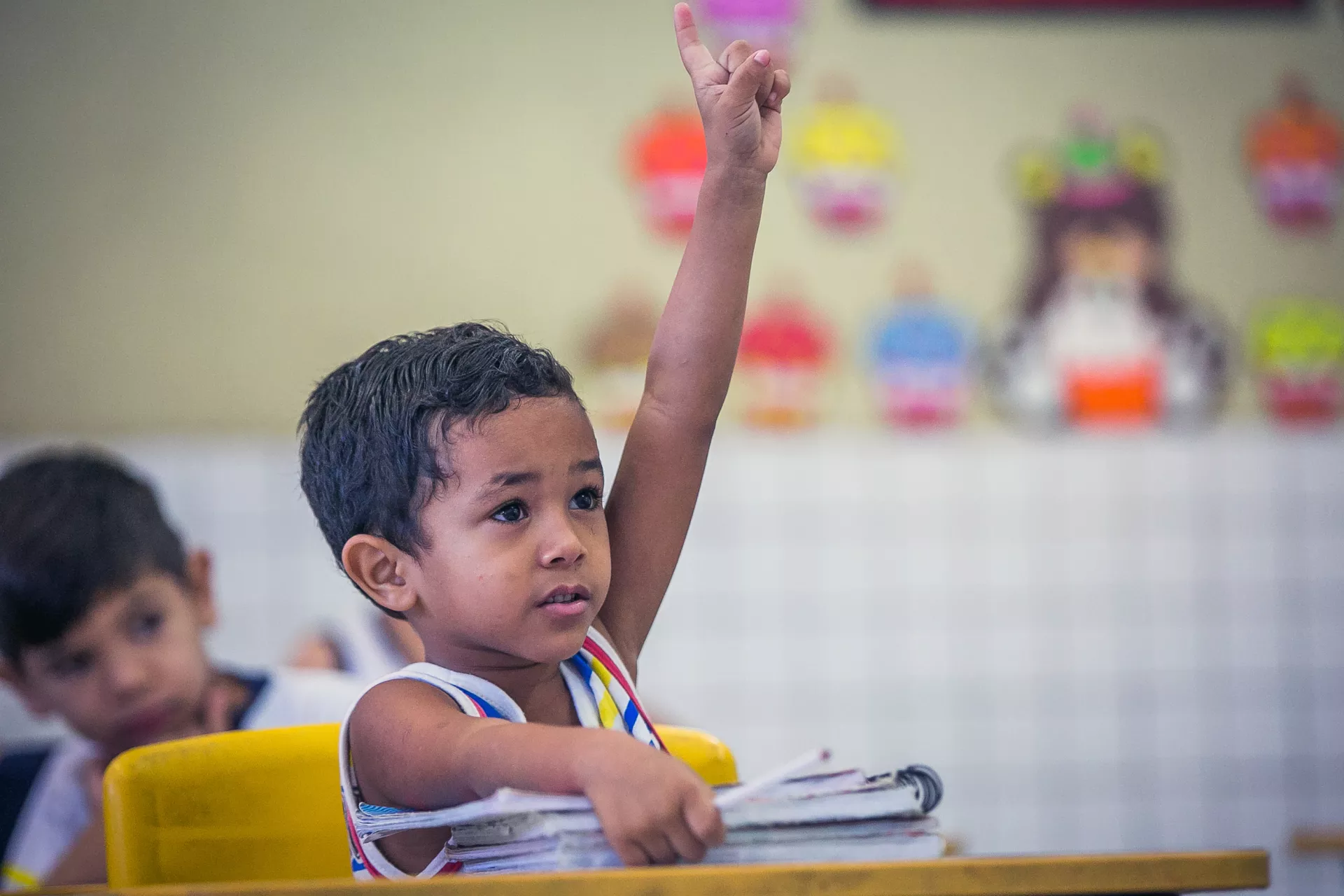 um menino pequeno, sentando em uma carteira escolar, levanta o bra&ccedil;o