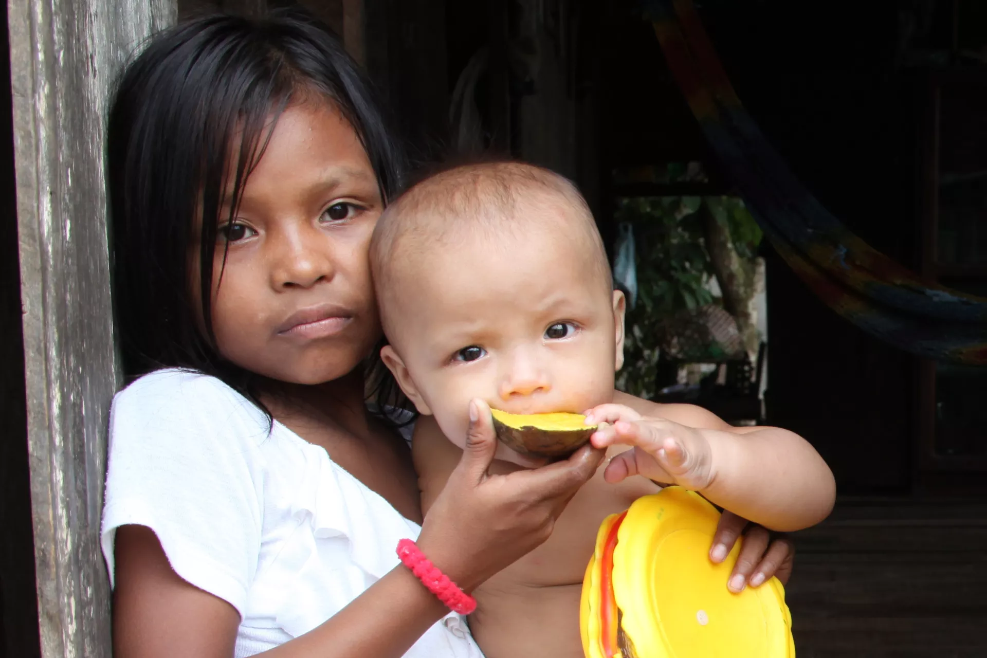 menina ind&iacute;gena ajuda beb&ecirc; ind&iacute;gena a comer uma manga