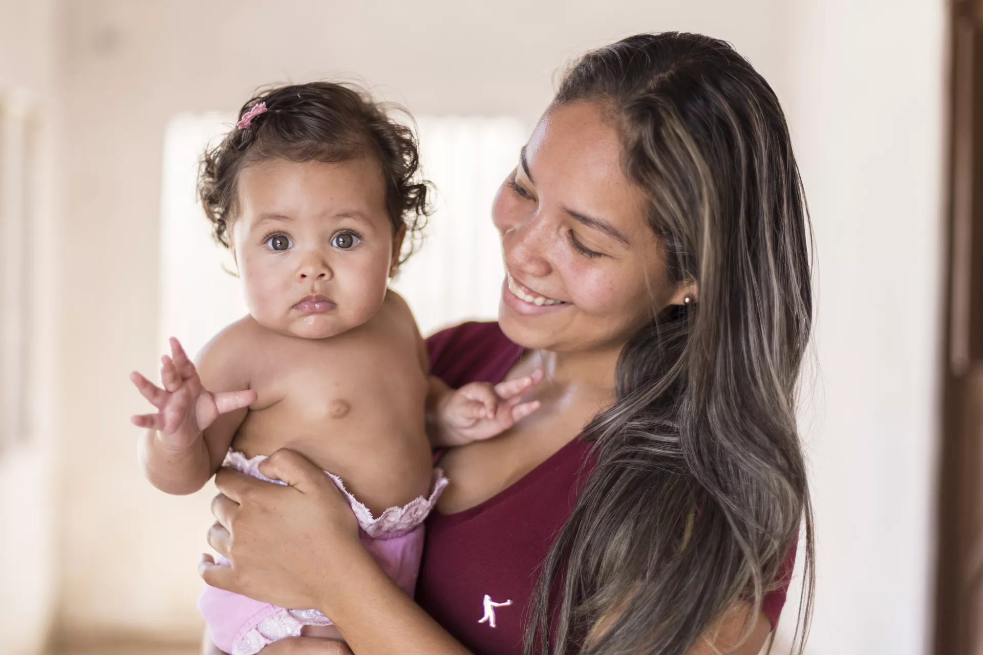 uma beb&ecirc; olha para a c&acirc;mera. Ela est&aacute; no colo da m&atilde;e. A m&atilde;e sorridente olha para a beb&ecirc;.