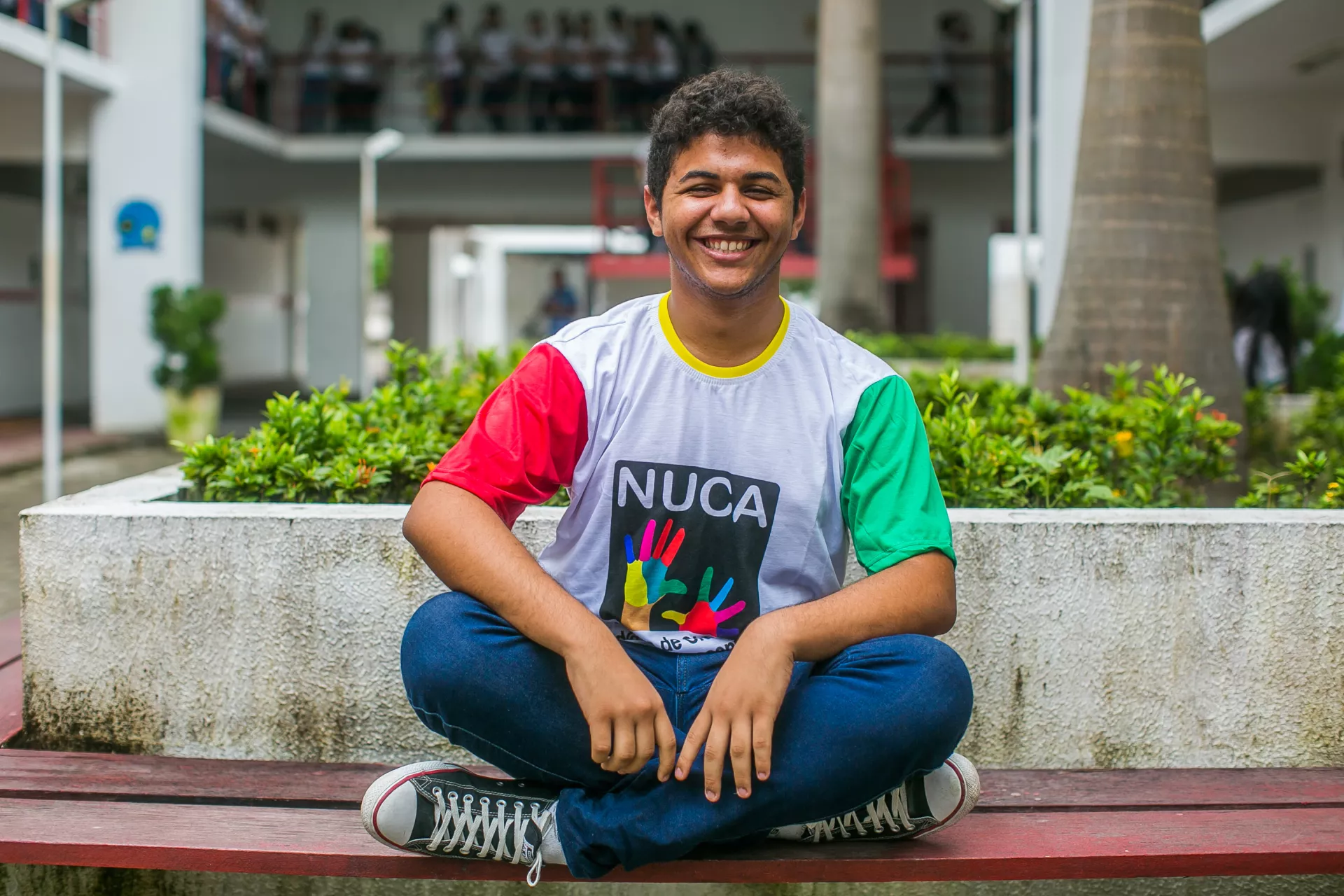 Felipe Caetano est&aacute; sentado no p&aacute;tio da escola. Ele usa a camiseta do NUCA, olha para a c&acirc;mera e sorri. Atr&aacute;s dele, com a imagem desfocada, est&aacute; a escola com diversos estudantes.