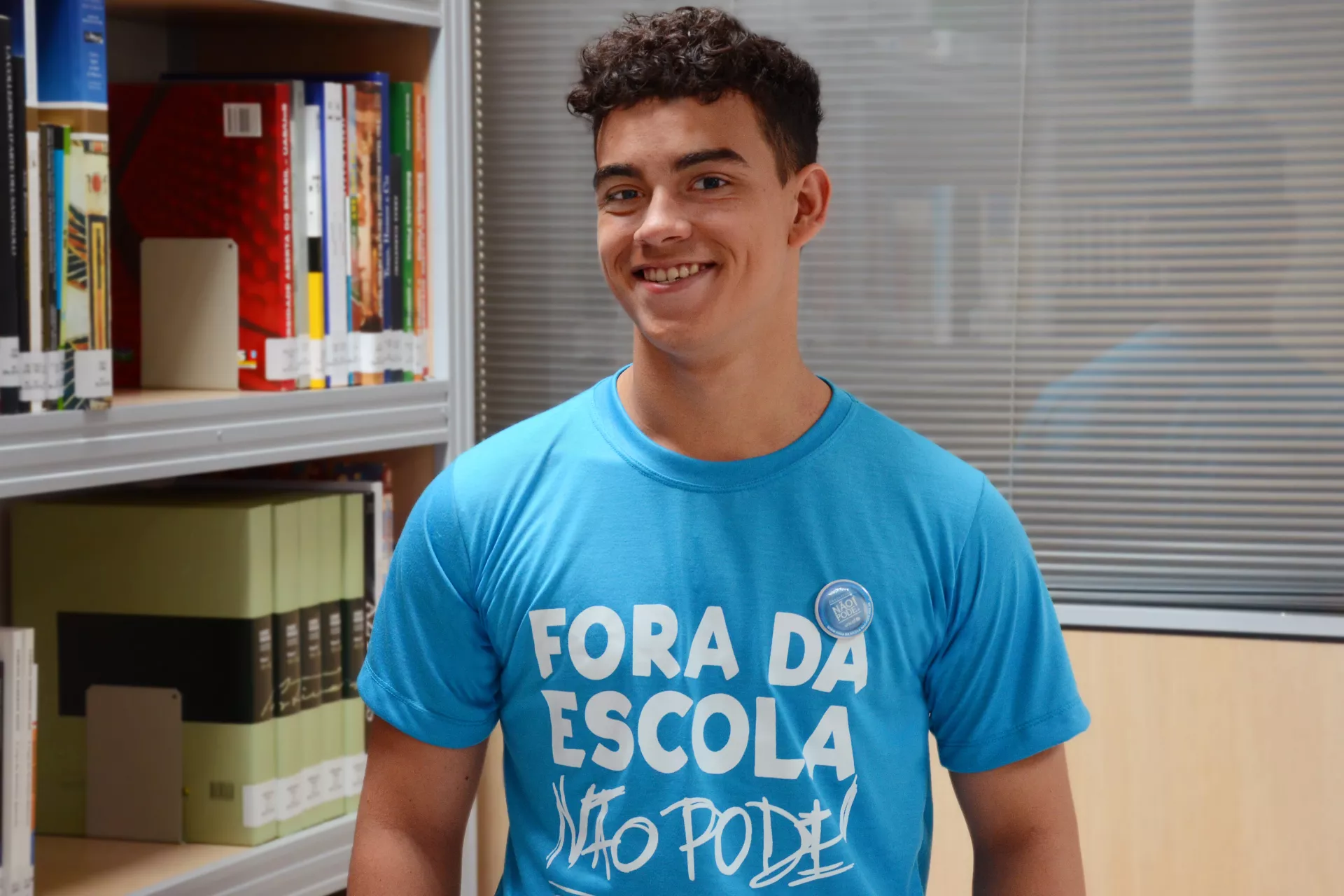 Adolescente com uma camiseta azul onde est&aacute; escrito 'fora da escola n&atilde;o pode' sorri para a c&acirc;mera. Ele est&aacute; ao lado de uma estante de publica&ccedil;&otilde;es e na frente de uma divis&oacute;ria de vidro.