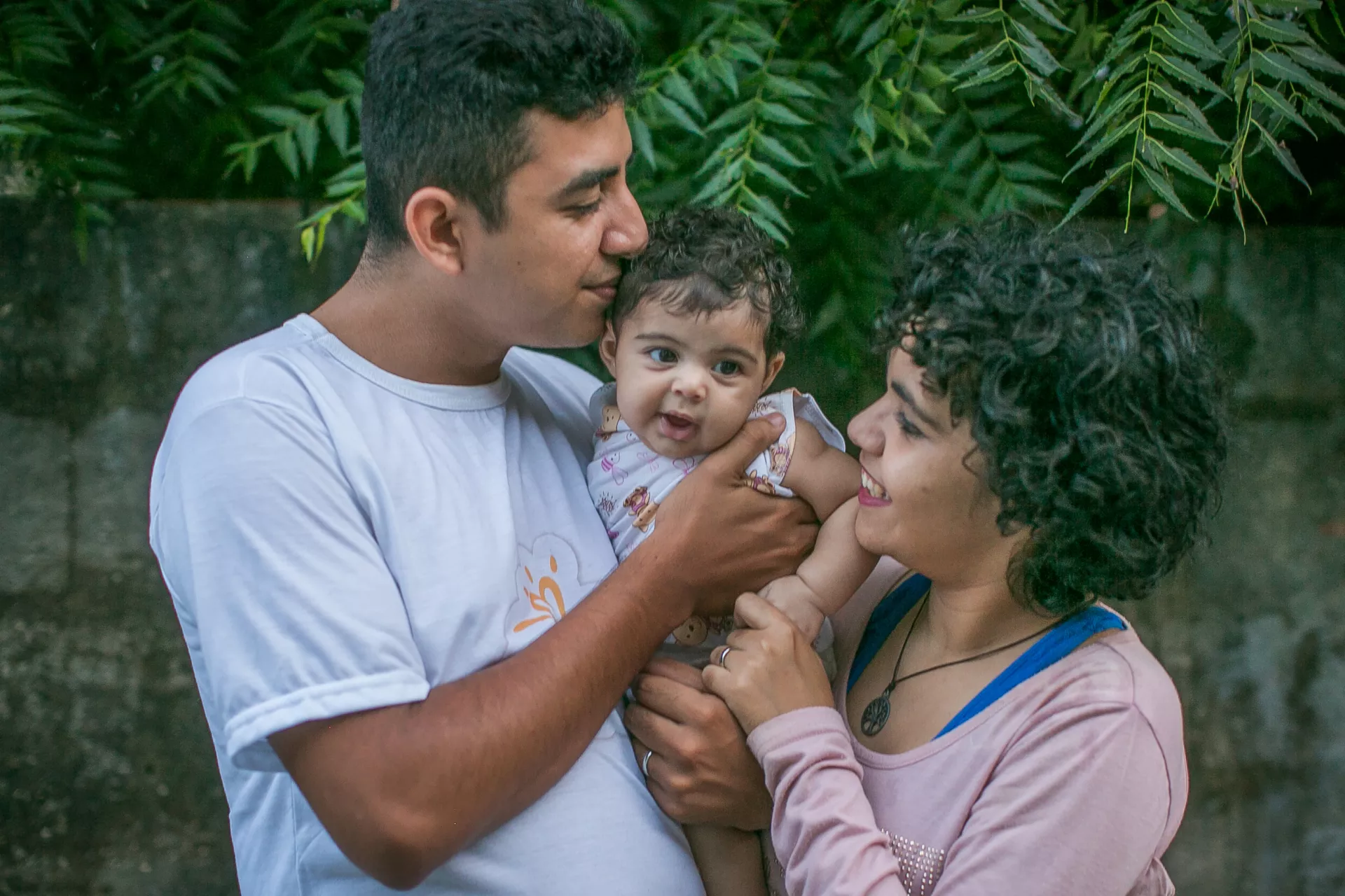 um pai e uma m&atilde;e seguram sua beb&ecirc; no colo. A beb&ecirc; est&aacute; olhando para a c&acirc;mera e sorrindo.