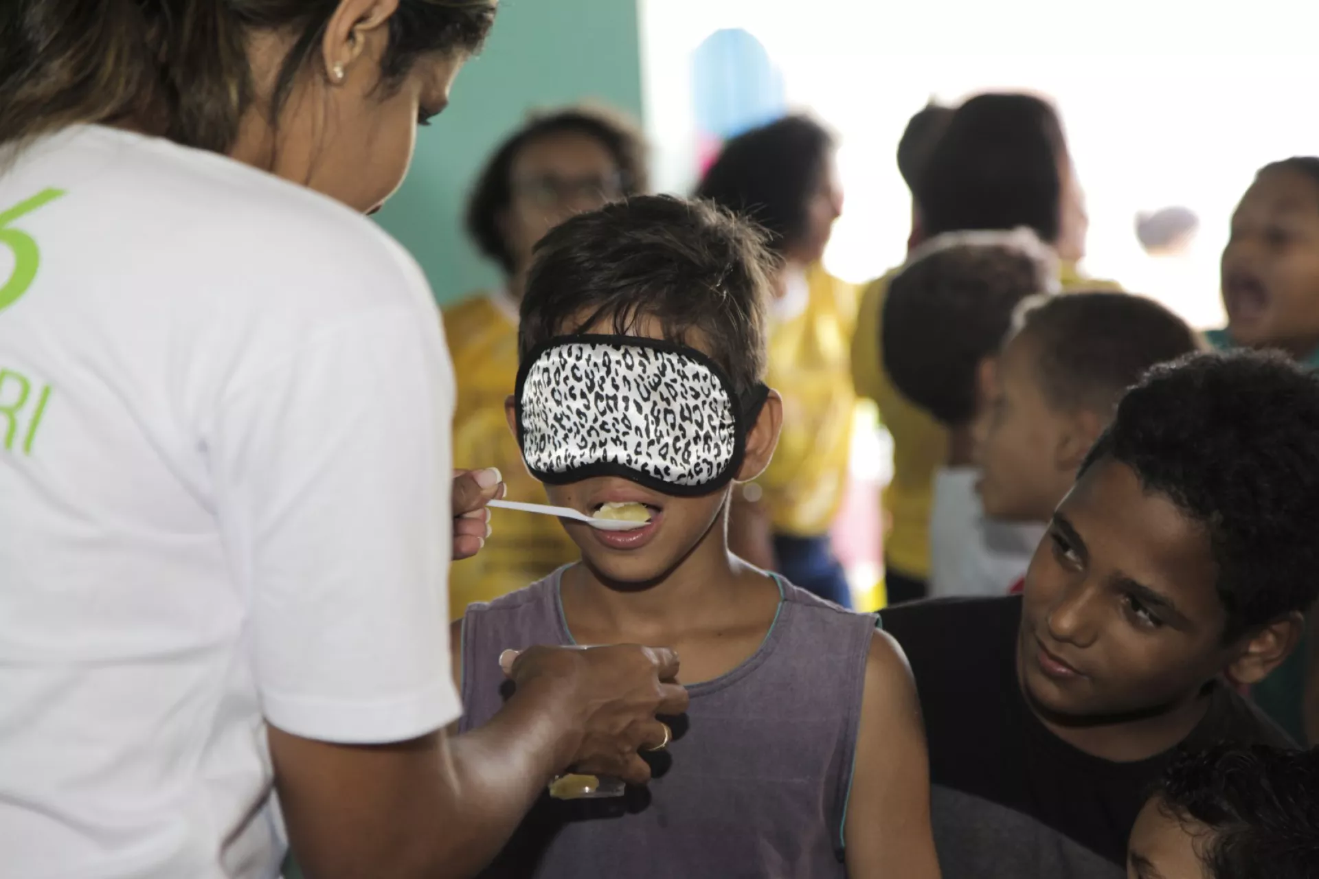 menino com olhos vendados prova uma fruta dada por uma profissional de saúde. Outras crianças estão em volta dele.