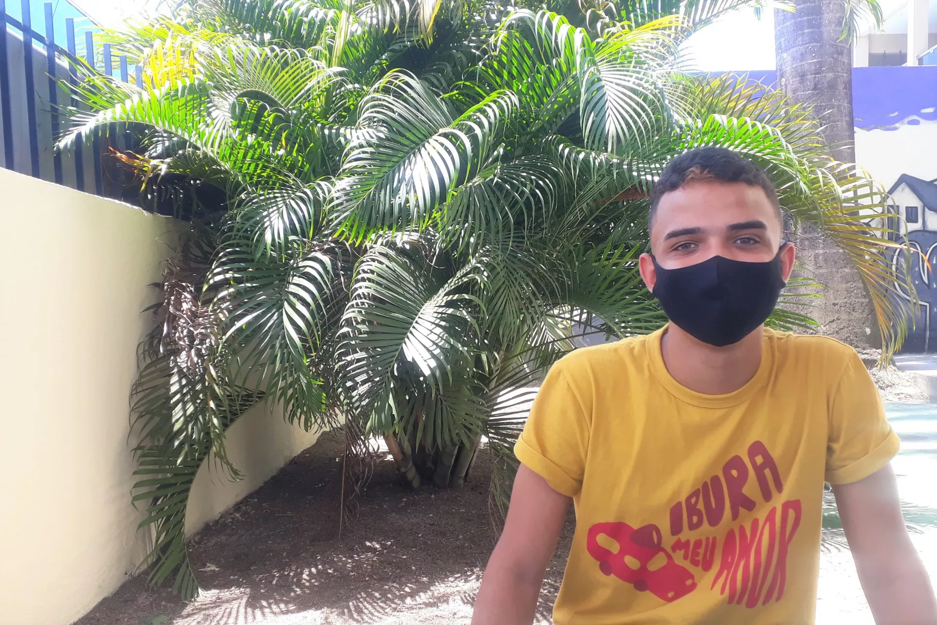 Foto mostra adolescente usando máscara e uma camiseta do projeto Ibura, Meu Amor. Ele está sentando ao ar livre, em frente a umas palmeiras.