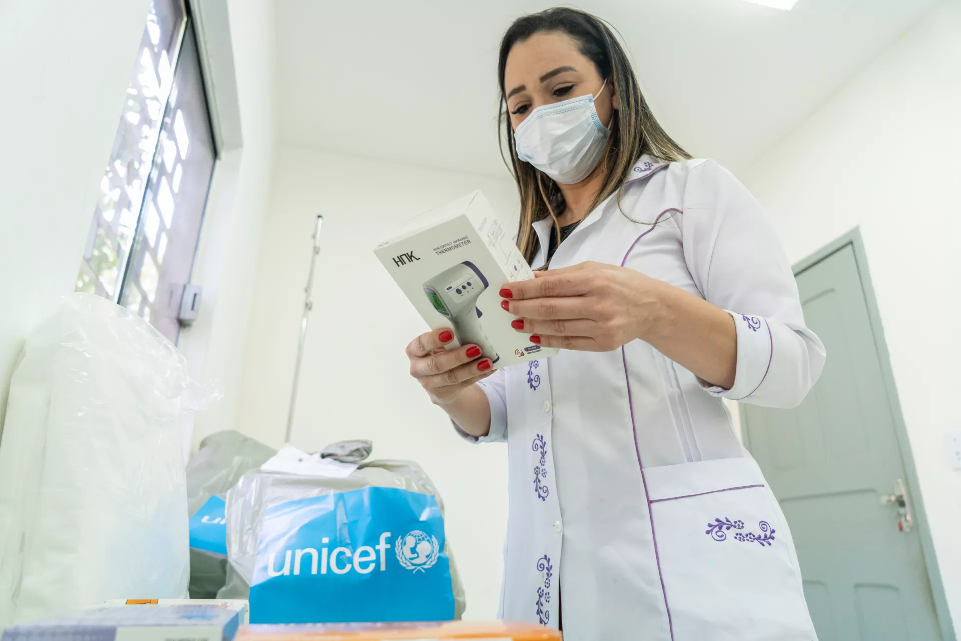 Foto mostra uma mulher vestindo um jaleco e usando m&aacute;scara segurando uma caixa de term&ocirc;metro. Atr&aacute;s dela vemos alguns sacos com o logo do UNICEF.