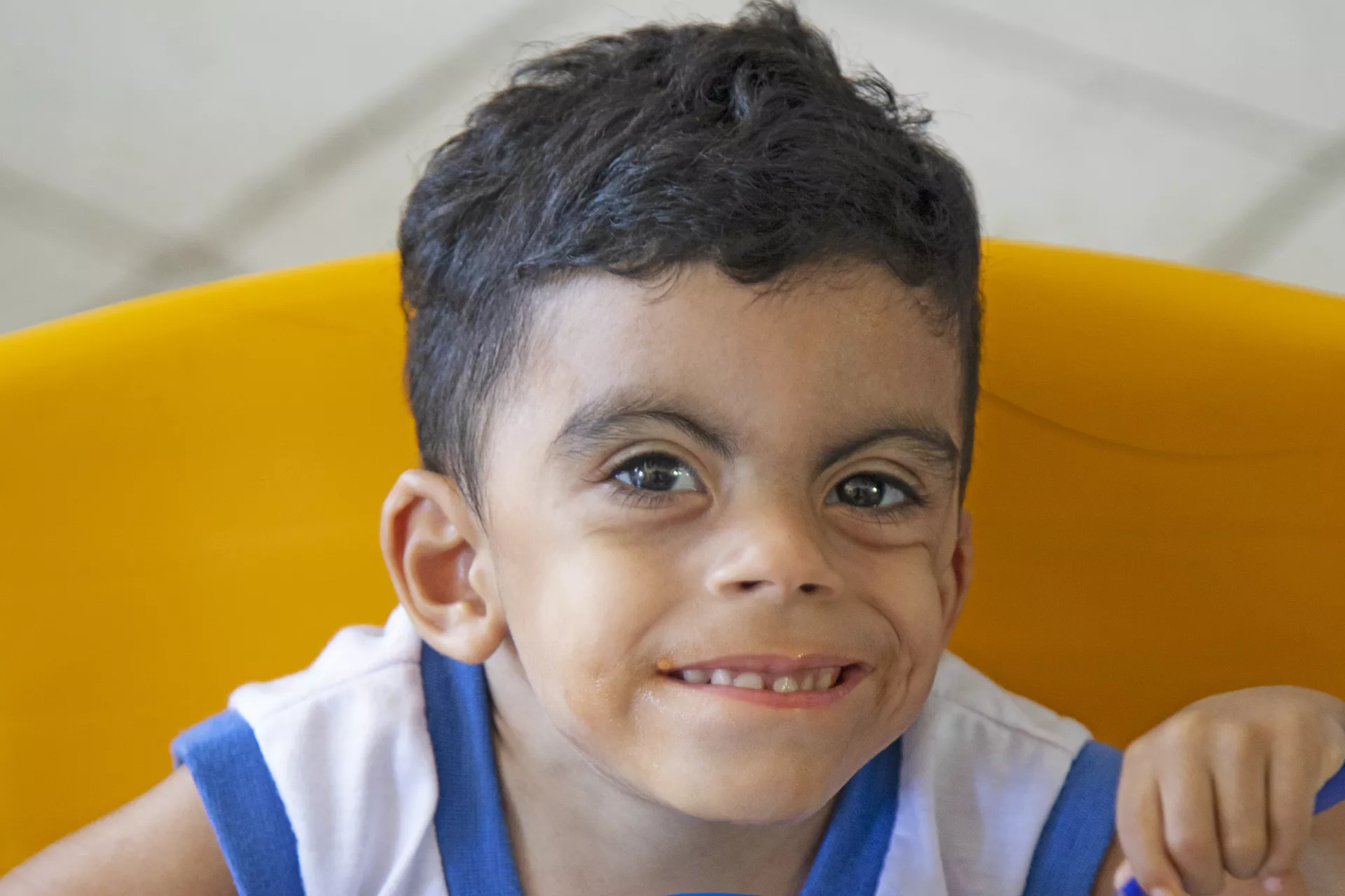 Foto mostra o rosto de um menino pequeno. Ele est&aacute; sorrindo e olhando para a c&acirc;mera.