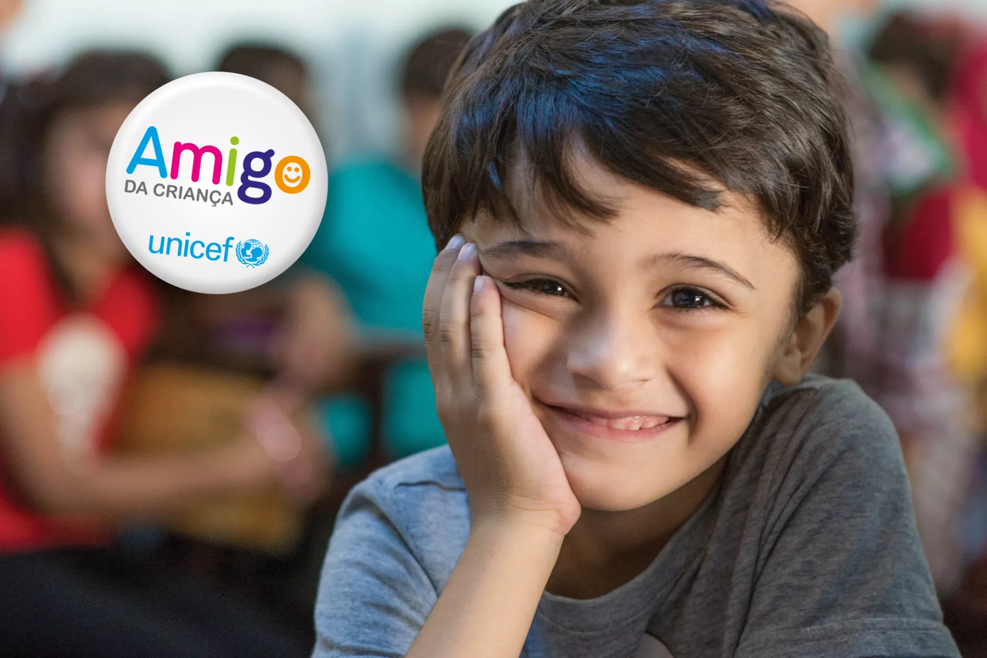 Foto mostra um menino sorridente, apoiando o rosto na m&atilde;o. Em cima da foto, do lado esquerdo, h&aacute; a marca do programa Amigo da Crian&ccedil;a, do UNICEF Brasil