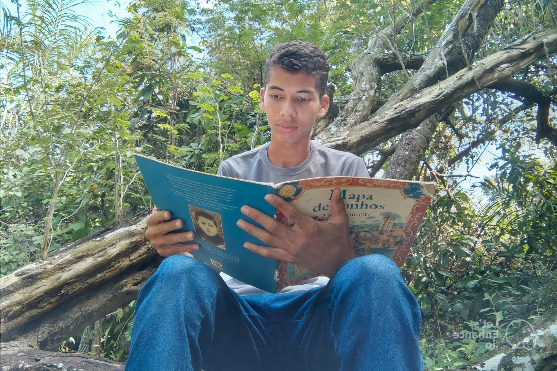 Foto mostra um adolescente lendo um livro. Ele est&aacute; sentado na frente de v&aacute;rias &aacute;rvores.