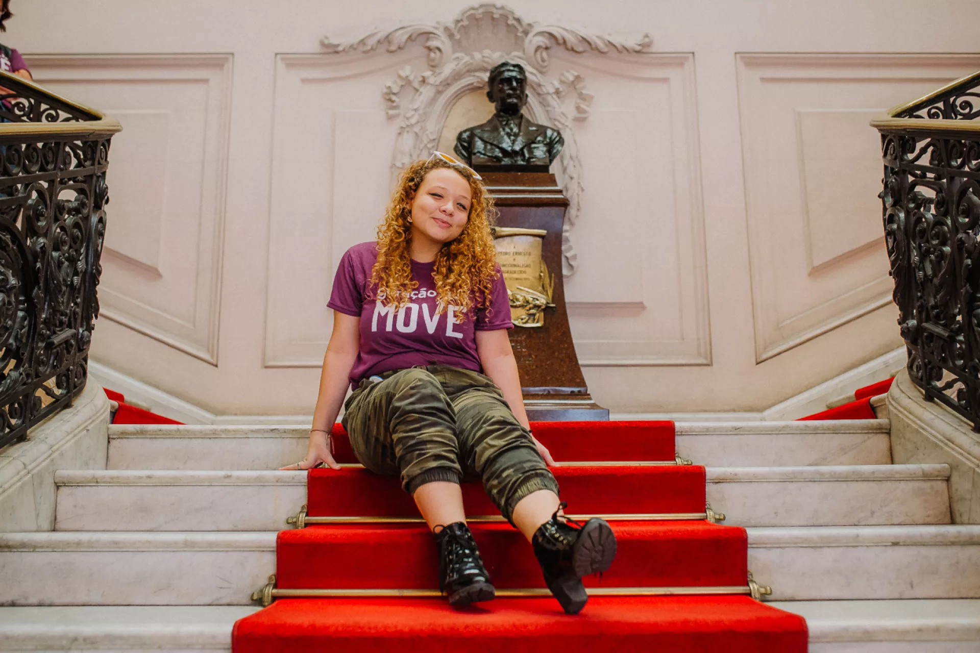 Foto mostra adolescente sentada em uma escadaria com tapete vermelho. Atrás dela há um busto de bronze de Pedro Ernesto, ex-prefeito da cidade do Rio de Janeiro, então Distrito Federal.