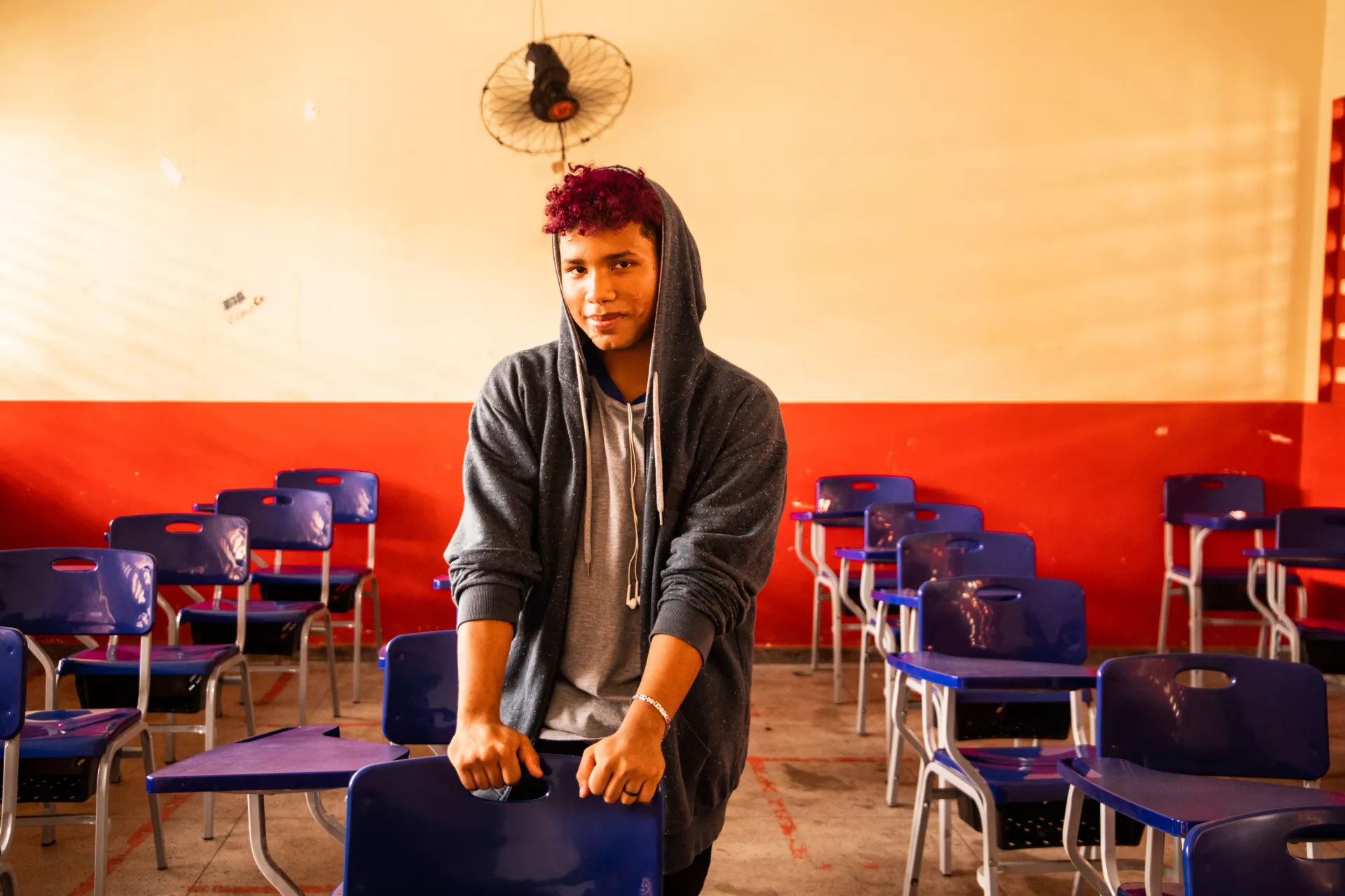 Foto mostra um adolescente em uma sala de aula vazia. Ele está de pé, apoiando-se em uma carteira escolar.
