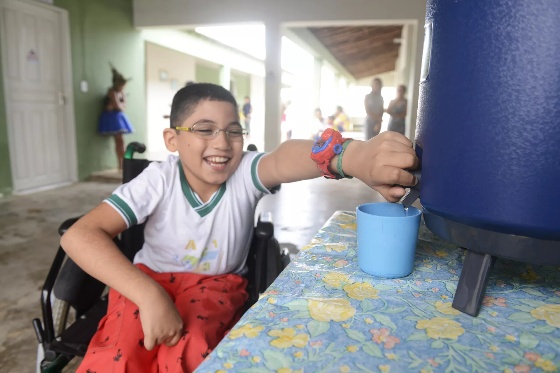 Foto mostra um menino pegando &aacute;gua de um filtro na escola. Ele est&aacute; sentado em uma cadeira de rodas