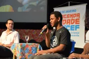 Foto mostra um adolescente falando em um microfone. Ele está sentado. Atrás dele aparece o banner da #AgendaCidadeUNICEF.
