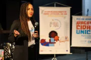 Foto mostra uma mulher falando no microfone. Ao lado dela, h&aacute; um banner da estrat&eacute;gia PIA