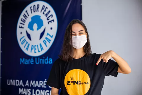 Foto mostra uma jovem em pé em frente a um banner da Luta pela Paz. Ela está usando a camiseta do projeto Zona Nossa e apontado o dedo para o logo que está na camiseta. Ela usa máscara.