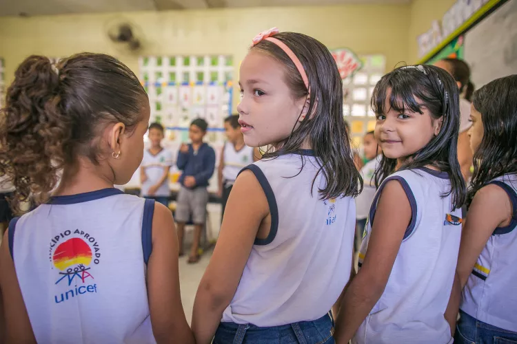 crian&ccedil;as est&atilde;o em roda na sala de aula. As tr&ecirc;s meninas em primeiro plano est&atilde;o de m&atilde;os dadas. Elas est&atilde;o de costas para a c&acirc;mera. A menina do meio olha pra o lado e a menina da direita olha para a c&acirc;mera. Nas costas do uniforme escolar est&aacute; o logo do Selo UNICEF.