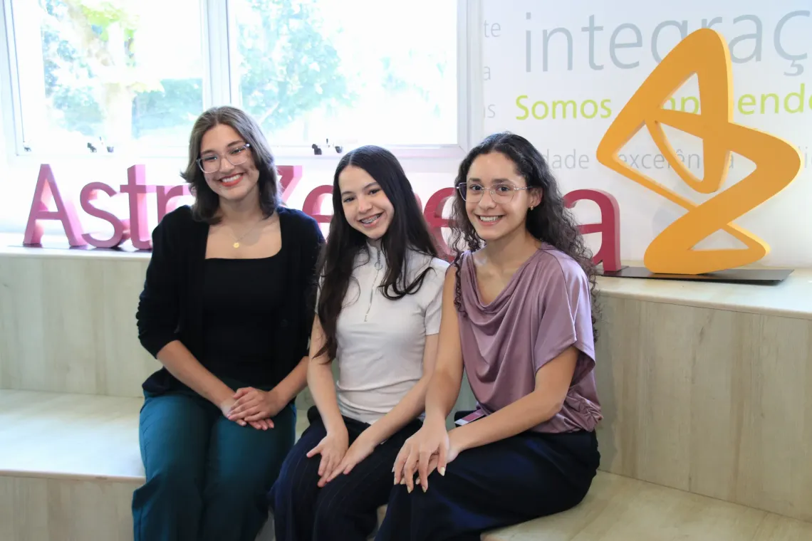 Mulher e duas jovens sentadas posam para foto em frente a um letreiro da AstraZeneca
