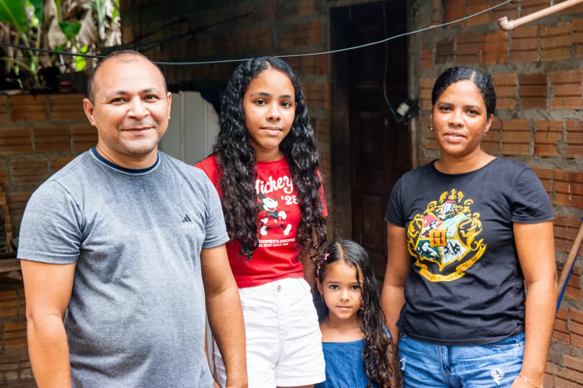 fam&iacute;lia de pai, m&atilde;e e duas filhas posam para foto em frente a uma casa simples
