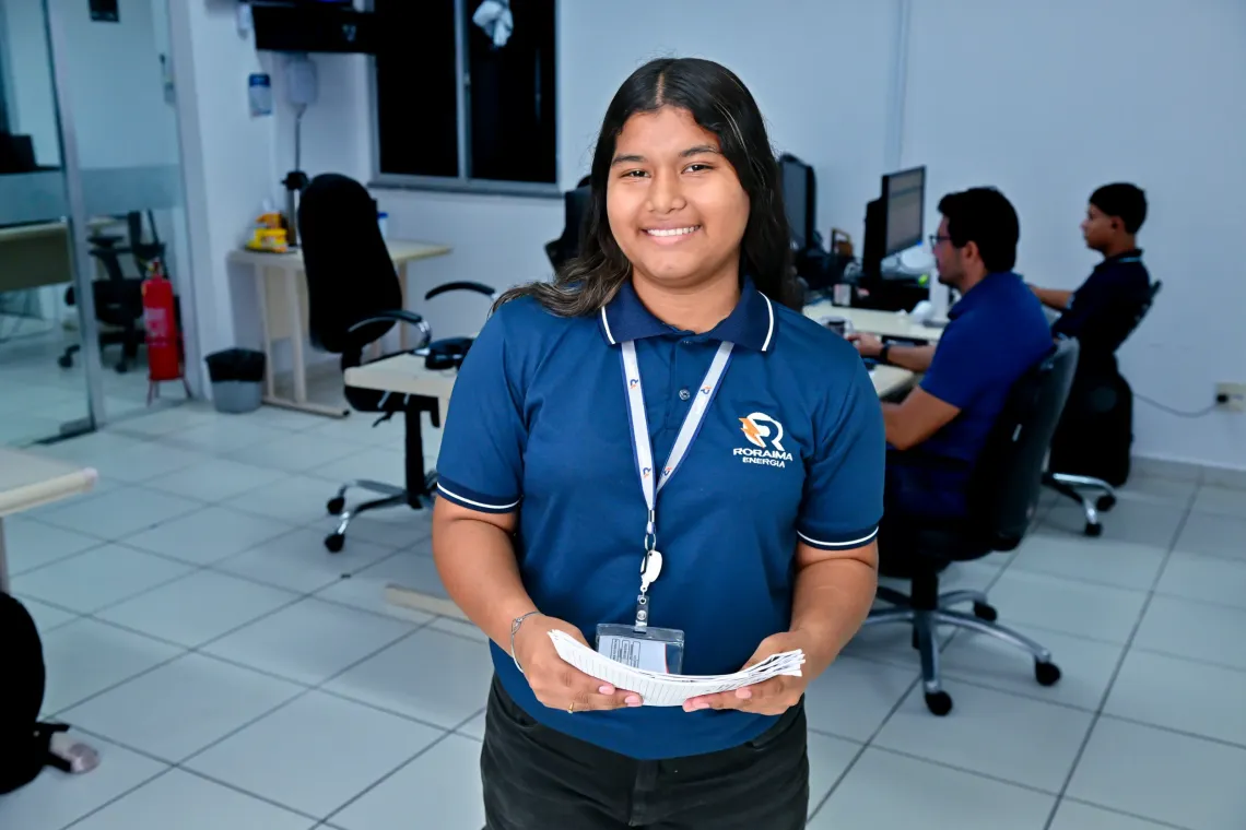 jovem de cabelos compridos e escuros vestindo uniforme de trabalho azul e crach&aacute; segura papeis nas m&atilde;os e posa para foto em um escrit&oacute;rio empresarial
