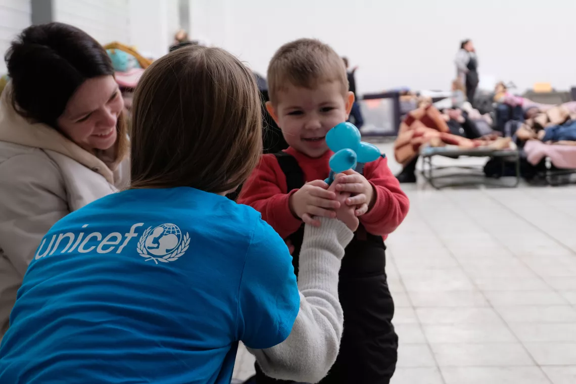 Foto mostra uma funcion&aacute;ria do UNICEF, de costas para a c&acirc;mera, vestindo a camiseta azul com o logo da institui&ccedil;&atilde;o. Ela est&aacute; entregando um bichinho feito de bal&atilde;o para um beb&ecirc;. 