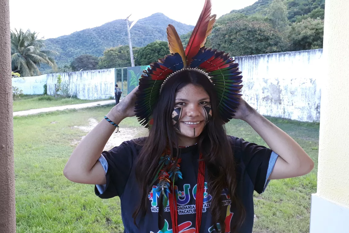 Foto mostra uma adolescente usando cocar e pinturas de povos tradicionais no rosto. Ela est&aacute; em um p&aacute;tio gramado. Ao fundo h&aacute; um muro e montanhas.