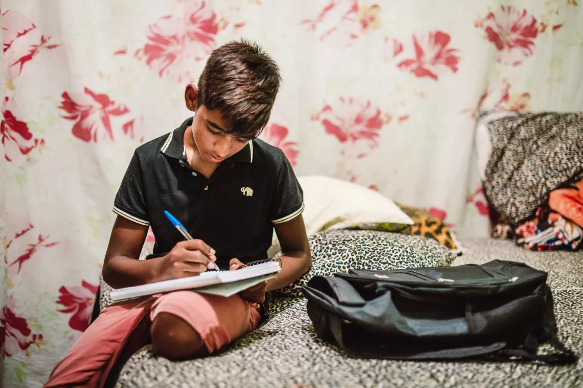 Foto mostra um menino sentado na cama escrevendo em um caderno. Do lado dele, em cima da cama, h&aacute; uma mochila.