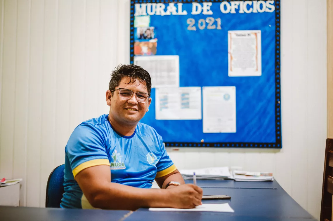 Foto mostra um homem sentado a uma mesa, em frente a um quadro de aviso. Ele est&aacute; olhando para a c&acirc;mera sorrindo.