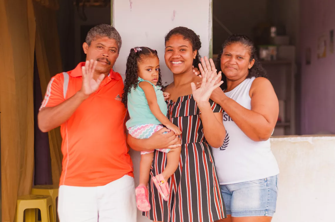 Foto mostra um home, uma adolescente com uma crian&ccedil;a pequena no colo, e uma mulher posando para a foto. Eles est&atilde;o encostados na parede externa de uma casa.