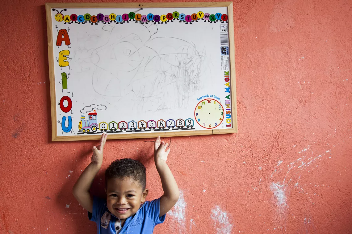 um menino pequeno est&aacute; em p&eacute;, encostado em uma parede. acima dele h&aacute; um quadro branco, com v&aacute;rias coisas escritas e rabiscadas.