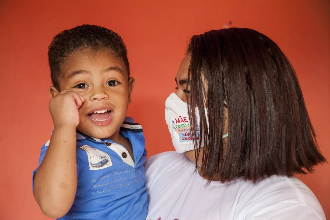 um menino pequeno est&aacute; no colo da m&atilde;e. ele olha para a c&acirc;mera sorrindo. a m&atilde;e est&aacute; de perfil, usando m&aacute;scara, e olha para o menino