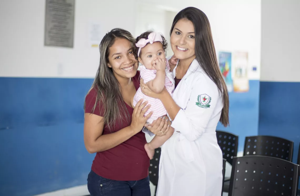 Uma m&atilde;e, uma beb&ecirc; e uma pediatra olham para a c&acirc;mera. A m&atilde;e e a pediatra est&atilde;o sorrindo.