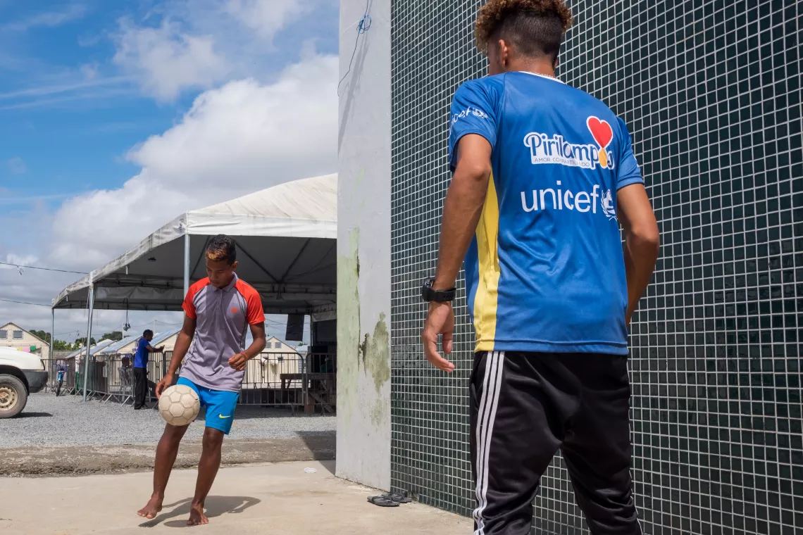 dois meninos jogam bola do lado de fora de um acampamento. Santiago est&aacute; de costas para a c&acirc;mera. Ele usa um colete azul com o logo do UNICEF e de seu parceiro Pirilampos