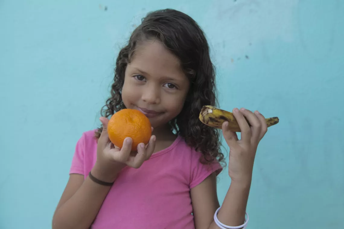 Mileena, 6 anos, olha para a câmera e sorri, enquanto segura uma laranja e uma banana. Ela veste uma camiseta rosa.