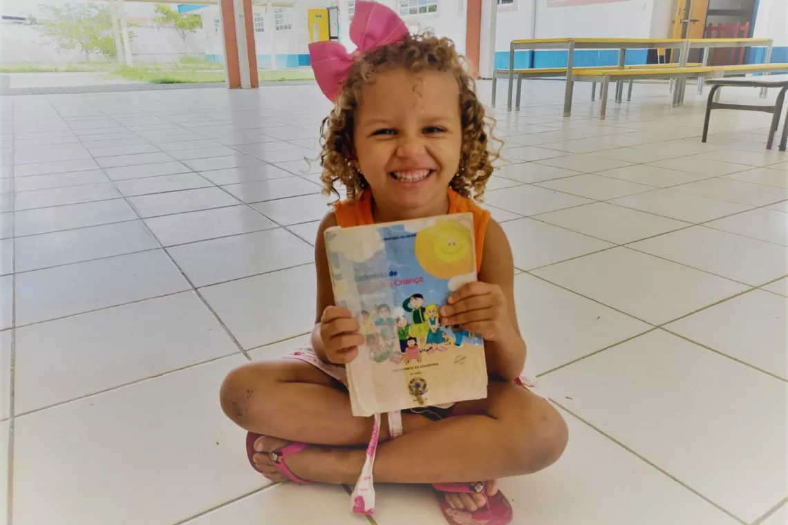 Foto mostra uma menina pequena sentada no ch&atilde;o, com as pernas cruzadas. Ela est&aacute; segurando e mostrando uma caderneta de vacina&ccedil;&atilde;o. Ela est&aacute; sorrindo.
