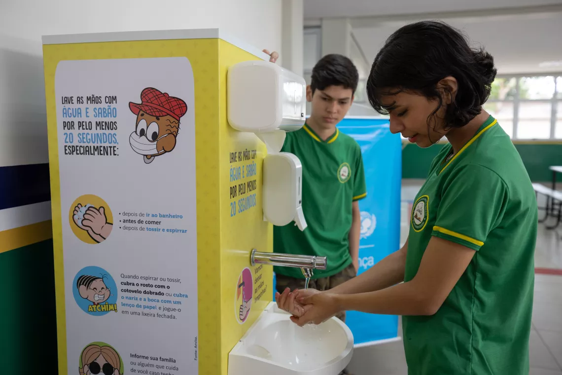 Foto mostra uma adolescente lavando as m&atilde;os em uma esta&ccedil;&atilde;o de lavagem de m&atilde;os que tem desenhos da Turma da M&ocirc;nica. Atr&aacute;s da esta&ccedil;&atilde;o, est&aacute; um adolescente, observando. Os dois est&atilde;o de uniforma escolar.